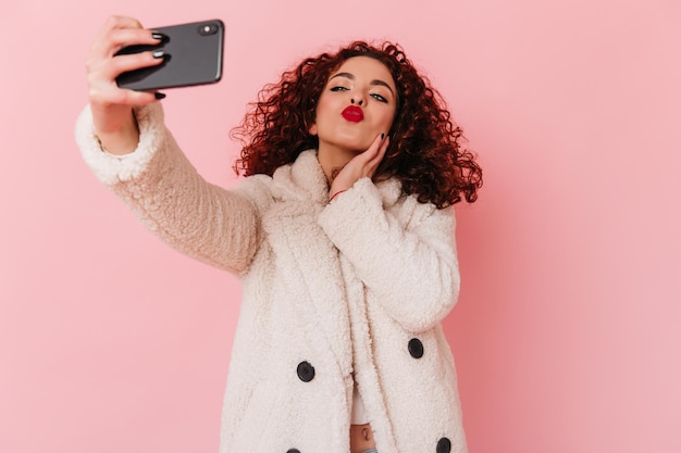 Free photo lady in white coat and cropped top blows kiss and takes selfie portrait of curly lady with bright lips on pink background