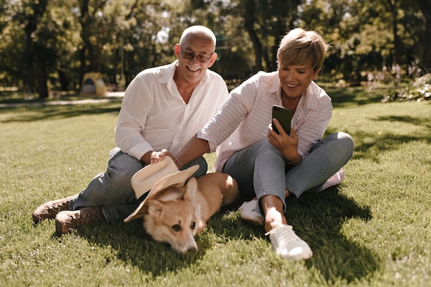 Lady with blonde hair in striped blouse and jeans making photo of dog and sitting on grass with old man in white shirt in park.