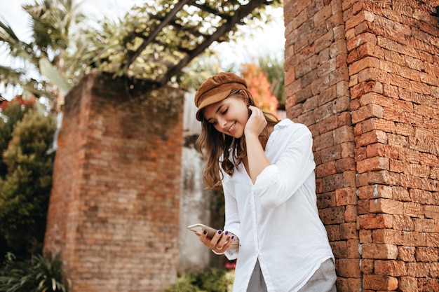 Lady with blue manicure holding smartphone. Girl in white blouse and gray pants posing near brick wall with tropical plants.