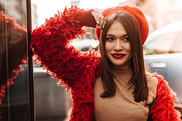 Free photo lady with dark straight hair and brown eyes dressed in red beret and eco-coat posing on street, leaning on glass wall.