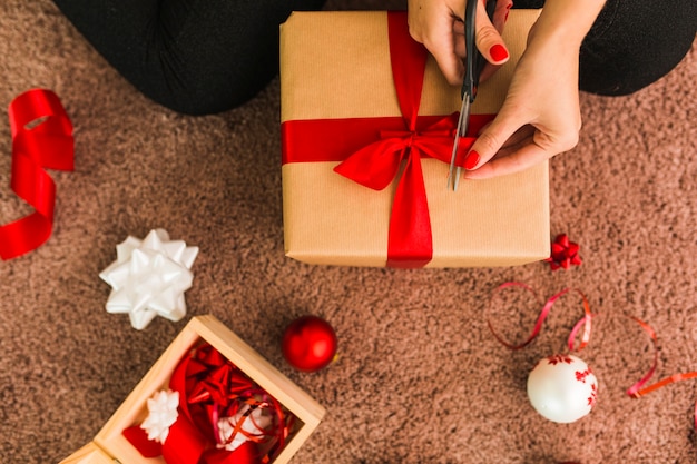 Lady with gift box and scissors near decorative bows, balls and ribbon on carpet