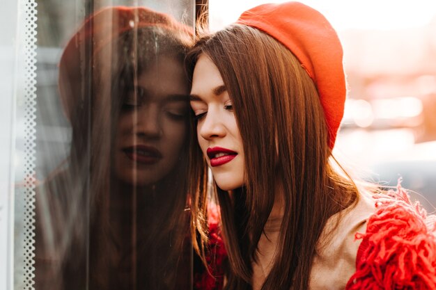 Lady with red lips in bright beret and sweater leaned on window, posing outside.