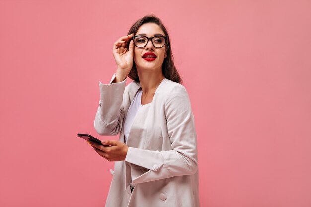 Lady with red lips dressed in cotton suit holds phone. Charming young woman with dark hair in glasses with black rim posing on camera.