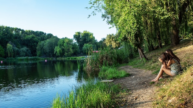 A lake with lots of green trees reflected in the water, two girls are sitting on the shore and reeds along it in Chisinau, Moldova