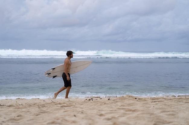 Free photo landscape image of male surfer busy walking on the beach at sunrise while carrying his surfboard under his arm with the ocean waves breaking in the background. young handsome male surfer on the ocean