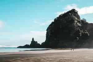 Free photo landscape of the piha beach and high rocks with the people walking around it under a blue sky