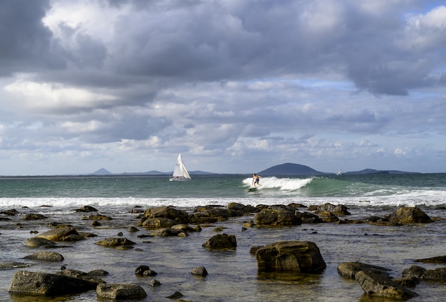 Free Photo landscape of the shore surrounded by the sea with ships and surfers on it under a cloudy sky