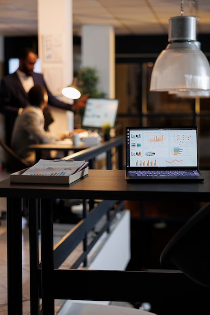 Free photo laptop computer with financial graphs on screen, standing on table in startup office, coworkers discussing company strategy in background. employees working late at night in startup office