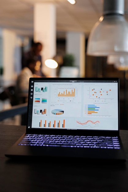 Laptop computer with marketing statistics graphs on screen, standing on table in startup office, coworkers discussing company strategy in background. Employees working overtime in startup office