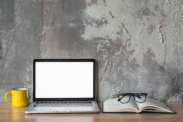 Free photo laptop; cup; book and spectacles on wooden desk in front of old wall