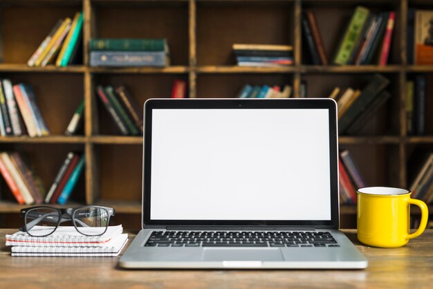 Laptop; cup; spectacles and spiral notepad on wooden desk