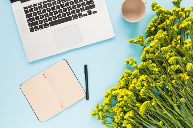 Laptop; diary; pen; empty ceramic cup and flower bouquet against blue background