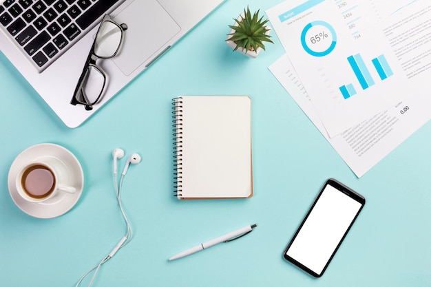 Laptop,eyeglasses,coffee cup,earphones,spiral notepad,pen,mobile phone and budget chart on office desk