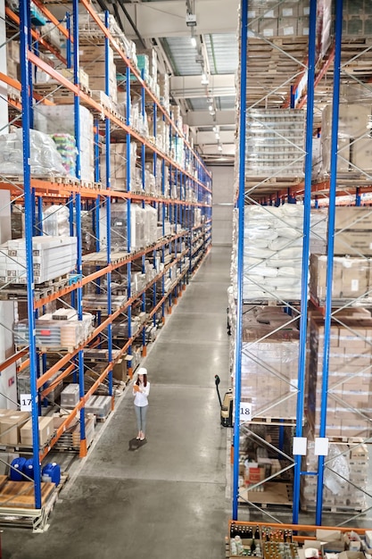 Free photo large warehouse. woman in white protective helmet with tablet standing in large industrial warehouse in aisle between high racks for goods