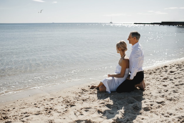 Free Photo lateral view of couple which is sitting on the sandy beach near the sea and looking on the breathtaking landscape