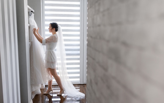 Free Photo lateral view of happy  bride in long veil who is preparing for the wedding day in room, dressing up in wedding dress