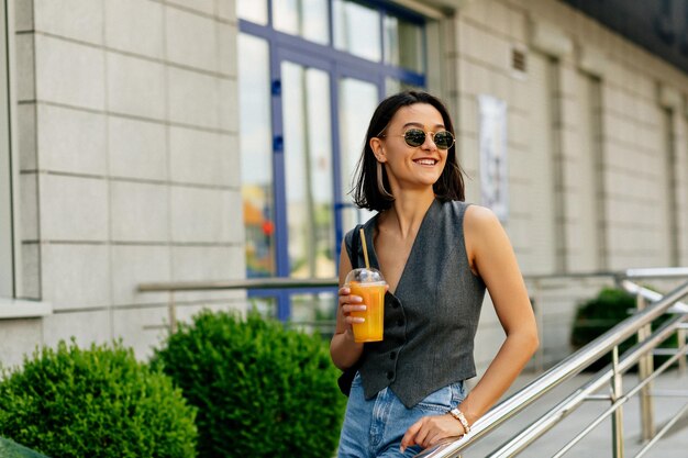 Laughing attractive stylish woman with short hairstyle is wearing grey jacket and jeans is drinking lemonade outdoor in warm sunny day