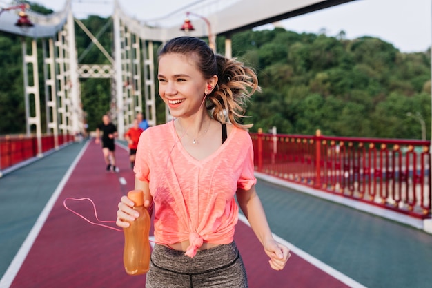 Laughing blonde girl holding bottle of juice and running down the cinder track. Amazing female model having fun at stadium.