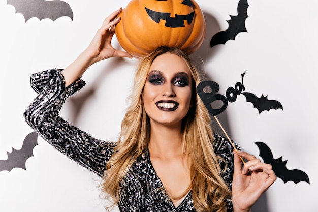 Laughing blonde witch posing with bats on wall. Magnificent young wizard holding big orange pumpkin.