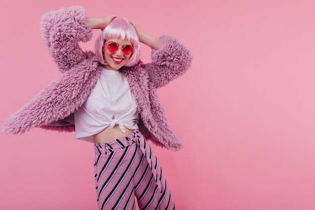 Free photo laughing dreamy girl in sunglasses touching her shiny periwig. smiling amazing young woman in fur jacket dancing on pink wall