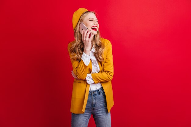 Laughing french young woman talking on phone.  adorable blonde girl with smartphone isolated on red wall.