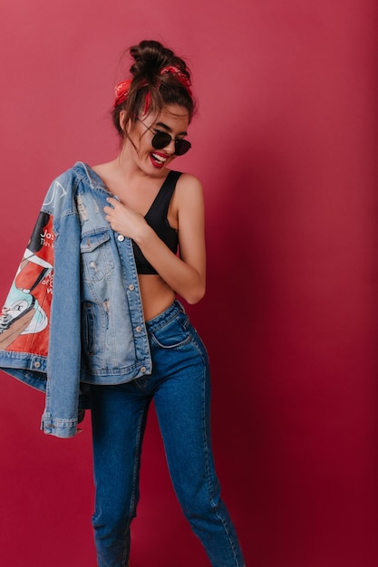 Laughing lightly-tanned girl with trendy makeup holding denim jacket while dancing