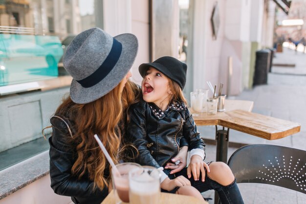 Laughing little girl wearing black hat and jacket resting on mom's knees and fooling around.