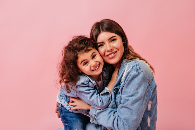 Laughing mother and daughter looking at camera. Front view of young woman with preteen child isolated on pink background.