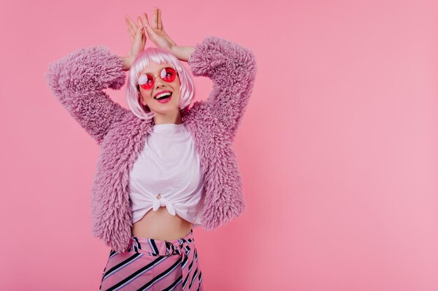 Laughing pleasant girl with tattoo posing in periwig on pink background Studio shot of ecstatic white female model wears elegant fur jacket and glasses