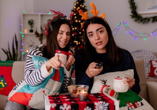 Laughing pretty young girl with holly wreath holds cup and points sitting on armchair with her friend holding tv remote and enjoying christmas time at home