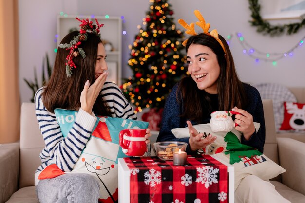 Laughing pretty young girls hold cups and look at each other sitting on armchairs and enjoying christmas time at home