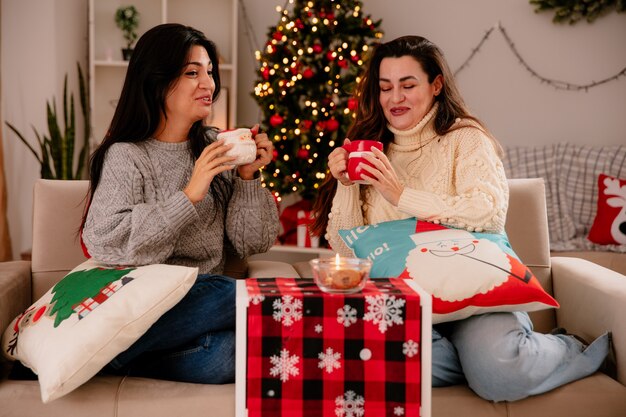 Laughing pretty young girls hold cups sitting on armchairs and enjoying christmas time at home