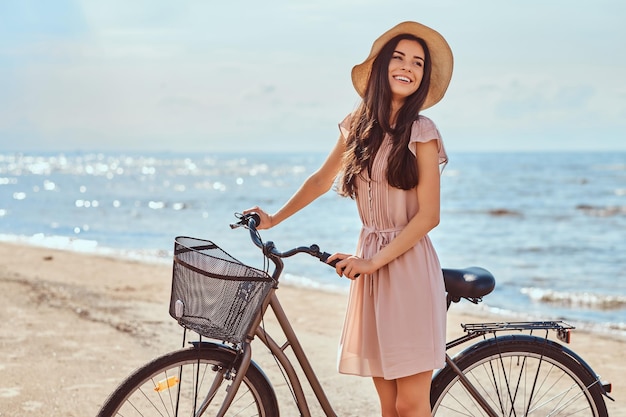 Laughing sensual girl with brown hair dressed in dress and hat posing with bicycle on the beach on a sunny day.