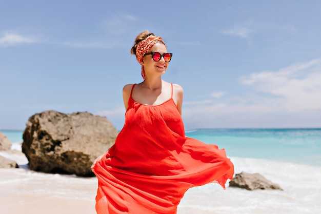 Laughing white girl fooling around at beach in sunny weekend. Outdoor photo of caucasian romantic lady in trendy dress dancing with rocks.