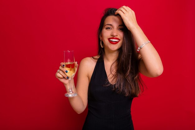 Laughing woman with adorable smile wearing black clothes with red lipstick holding a glass of champagne posing over red wall