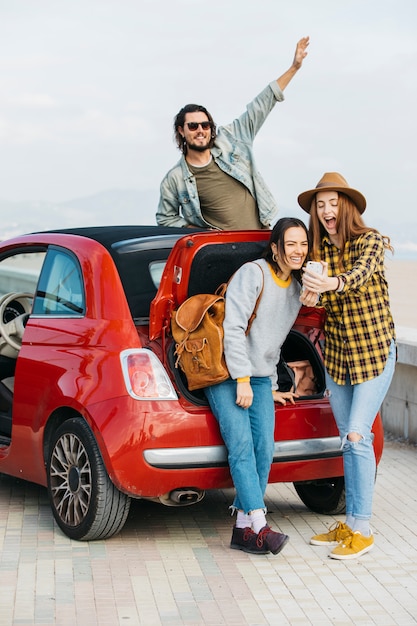 Laughing women taking selfie on smartphone near car trunk and man leaning out from auto