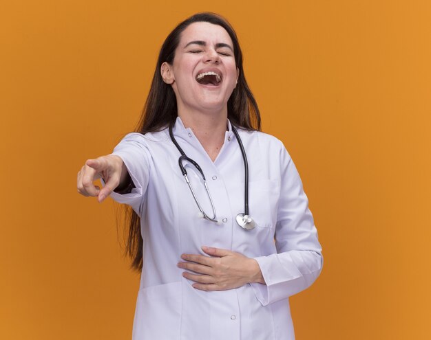 Laughing young female doctor wearing medical robe with stethoscope puts hand on belly and points forward isolated on orange wall with copy space