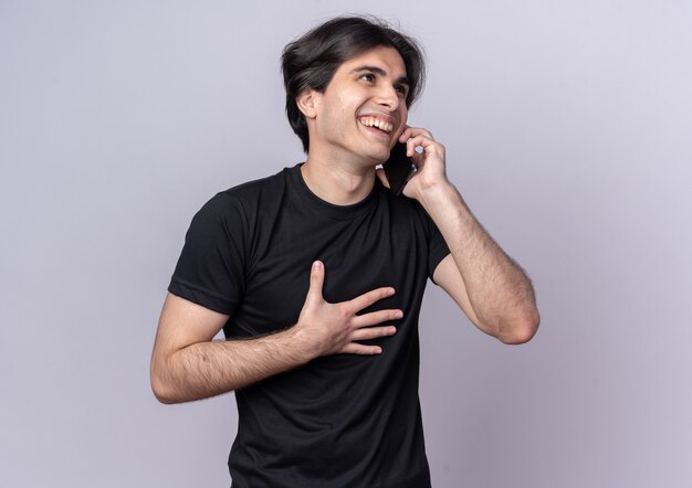 Laughing young handsome guy wearing black t-shirt speaks on phone putting hand on chest isolated on white wall