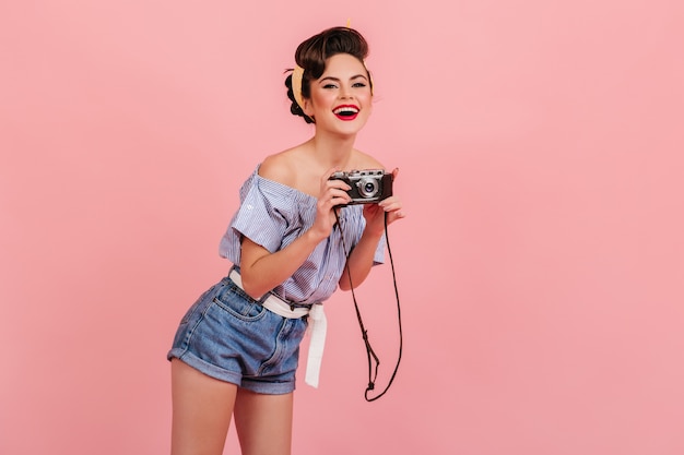 Laughing young woman in denim shorts taking pictures. Studio shot of pinup girl with camera isolated on pink background.