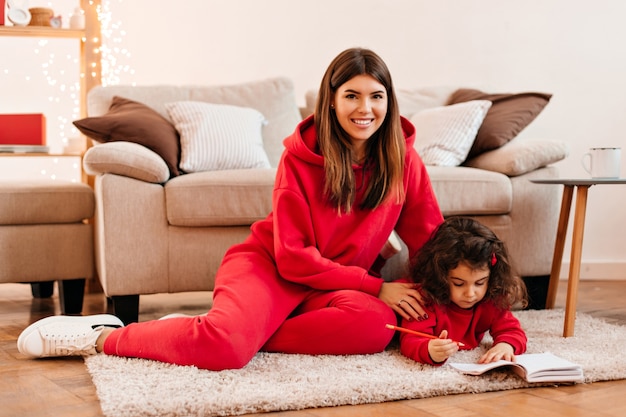 Laughing young woman teaching daughter to write. Indoor shot of happy mother and kid sitting on carpet.