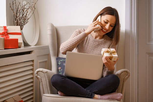 Laughing young woman with laptop talking on mobile phone while sitting in a comfortable armchair