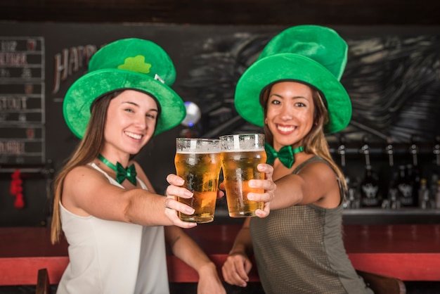 Free photo laughing young women in saint patricks hats showing glasses of drink at bar counter