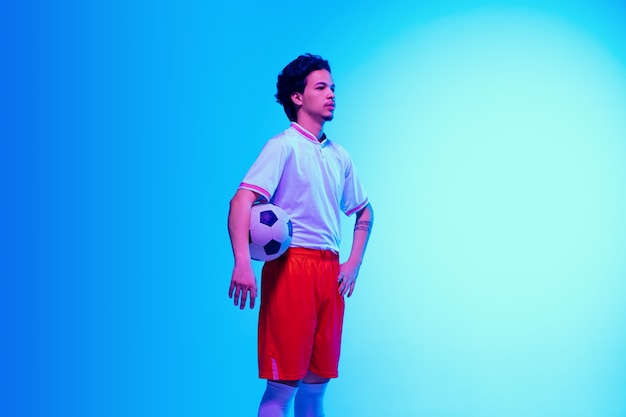 Leader. Football or soccer player on gradient blue studio wall in neon light - posing confident with ball. Copyspace.