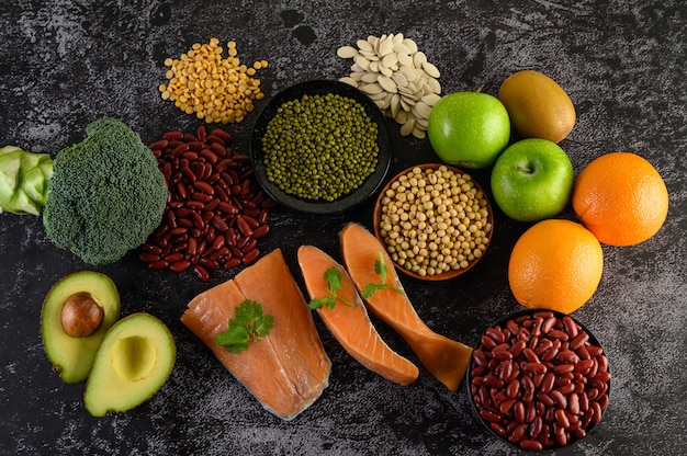 Legumes, broccoli, fruit, and salmon placed on a black cement floor.