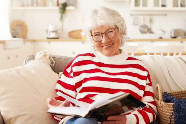 Leisure, self education, hobby and retirement concept. Picture of good looking mature senior female in striped sweater and stylish eyewear enjoying reading in living room, smiling joyfully