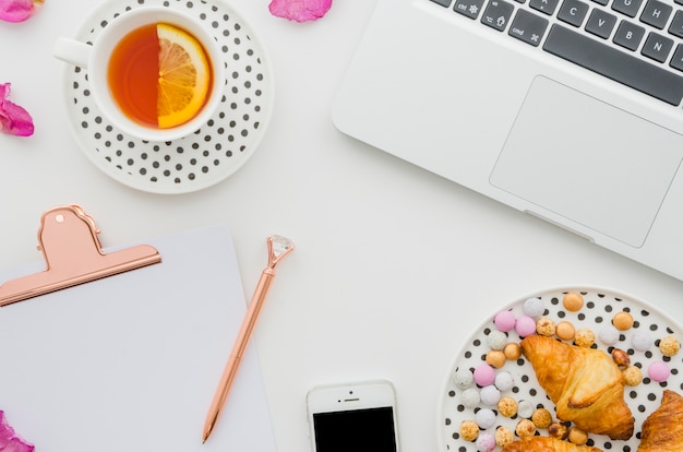 Lemon tea cup; laptop; croissant; candies; mobile phone; pen and clipboard on white backdrop