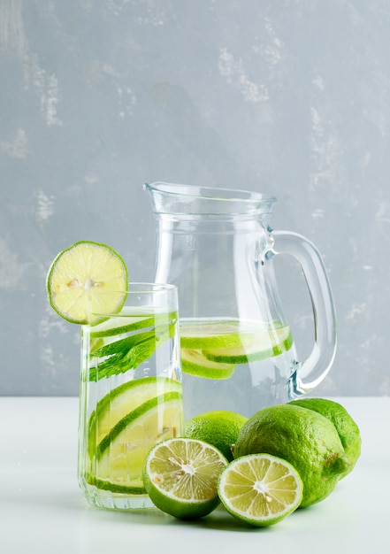 Free photo lemonade in glass and jug with lemon, basil side view on white and plaster