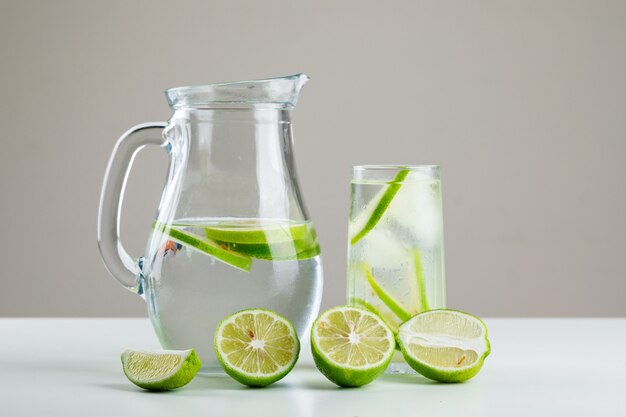 Lemonade in glass and jug with lemons side view on white and grey