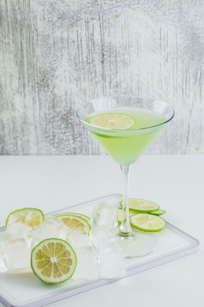 Free photo lemonade in a glass with lemon, cutting board, ice cubes side view on white and grungy