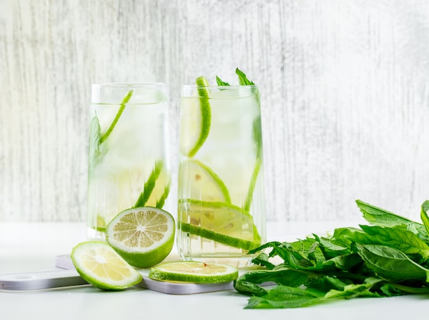 Free photo lemonade in glasses with lemon, basil, cutting board side view on white and grungy